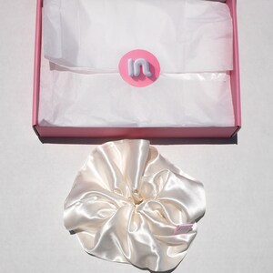 May include: A satin ivory scrunchie sits next to a pink box with white tissue paper. The box has a pink and white logo. The scrunchie is made of a shiny, silky material. The box is open, revealing the contents.