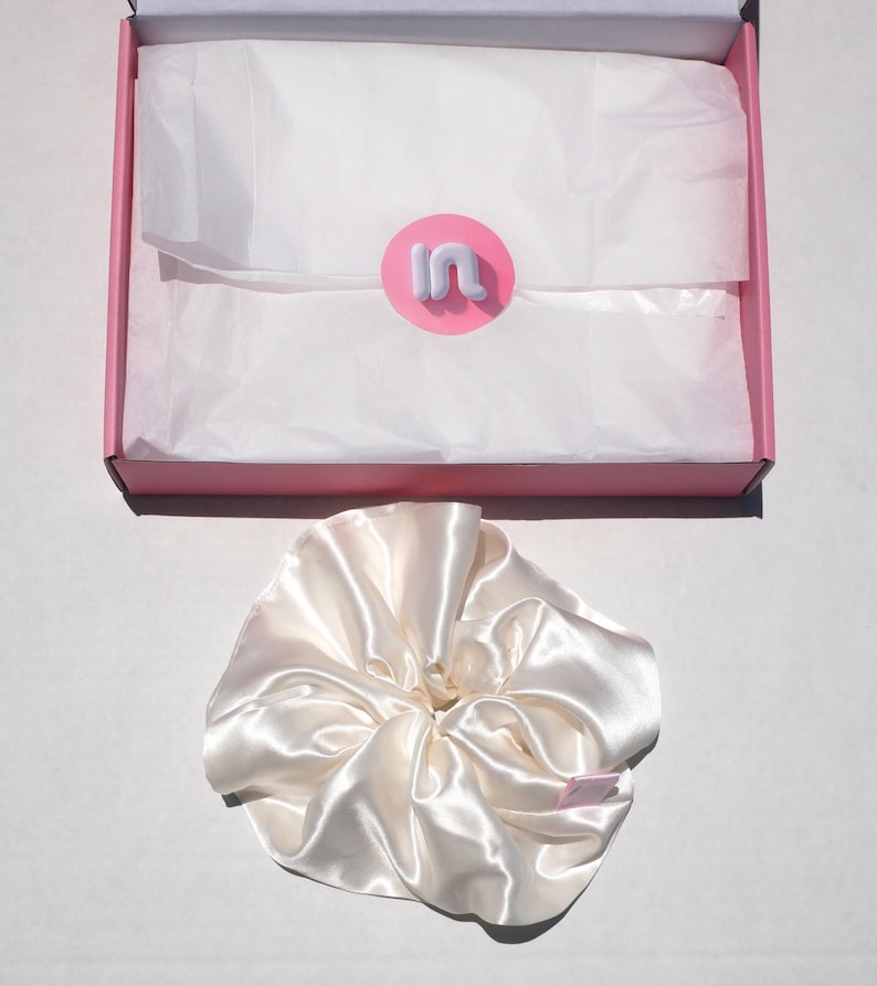 May include: A satin ivory scrunchie sits next to a pink box with white tissue paper. The box has a pink and white logo. The scrunchie is made of a shiny, silky material. The box is open, revealing the contents.