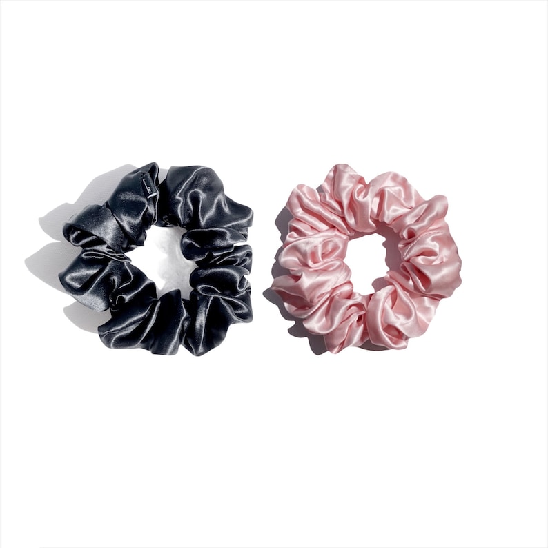 May include: Two satin scrunchies, one in a deep charcoal gray and the other in a soft blush pink, are displayed against a white background. The scrunchies are round and gathered, showcasing a shiny, smooth texture.