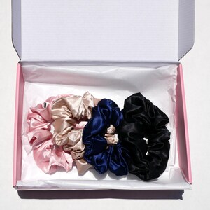 May include: A set of four satin scrunchies in a pink box. The scrunchies are in shades of pink, beige, navy blue, and black. The box is pink with a white interior.