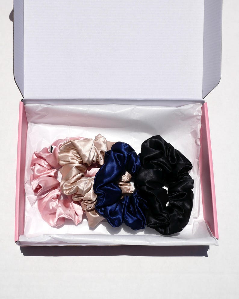 May include: A set of four satin scrunchies in a pink box. The scrunchies are in shades of pink, beige, navy blue, and black. The box is pink with a white interior.