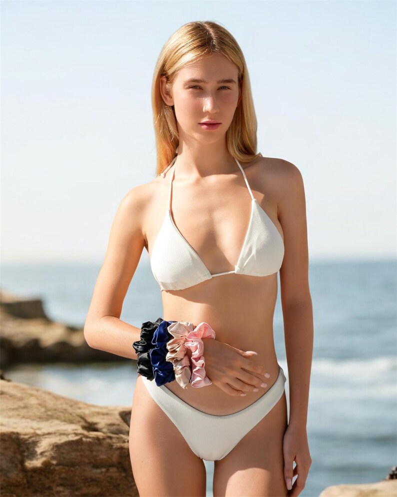May include: A woman wearing a white bikini with a selection of silk scrunchies in black, navy blue, and shades of pink and beige. The scrunchies are on the woman's wrist. The background is a beach scene with rocks and the ocean.
