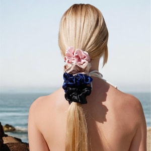 May include: A person's blonde hair is styled in a ponytail, secured with a stack of four satin scrunchies. The scrunchies are in shades of pink, tan, blue, and black. The background shows a beach and ocean.