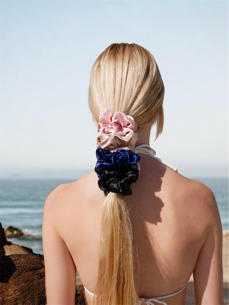May include: A person's blonde hair is styled in a ponytail, secured with a stack of four satin scrunchies. The scrunchies are in shades of pink, tan, blue, and black. The background shows a beach and ocean.
