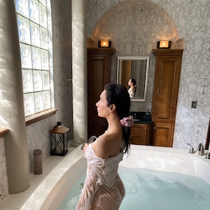 May include: A person in a white, wet shirt stands in a white bathtub filled with water. The bathroom has a large window with glass blocks, a wooden cabinet, and a mirror. A pink scrunchie is in the person's hair.