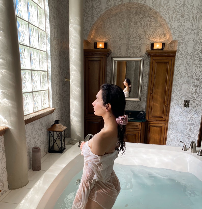 May include: A person in a white, wet shirt stands in a white bathtub filled with water. The bathroom has a large window with glass blocks, a wooden cabinet, and a mirror. A pink scrunchie is in the person's hair.