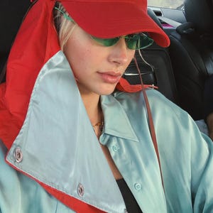 May include: A bright red baseball cap with a long fabric neck flap, paired with narrow green-tinted sunglasses and a light blue satin button-down shirt featuring a large, folded collar with silver-toned metal snap buttons along the front edge.