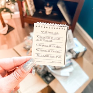 May include: A small notepad with a spiral binding, featuring the text "PCS Prep List." The list includes instructions such as "Rummage through all of the shit" and "Purge all of the shit." The notepad is held in front of a blurred background.
