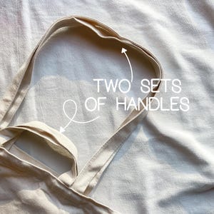 May include: A beige canvas tote bag with two sets of handles. The bag is folded in half and the handles are visible.
