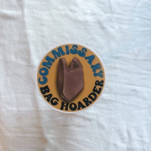 Op de afbeelding: Een ronde sticker met een gele achtergrond en blauwe tekst die "Commissary Bag Hoarder" luidt, met een bruine plastic tas in het midden.