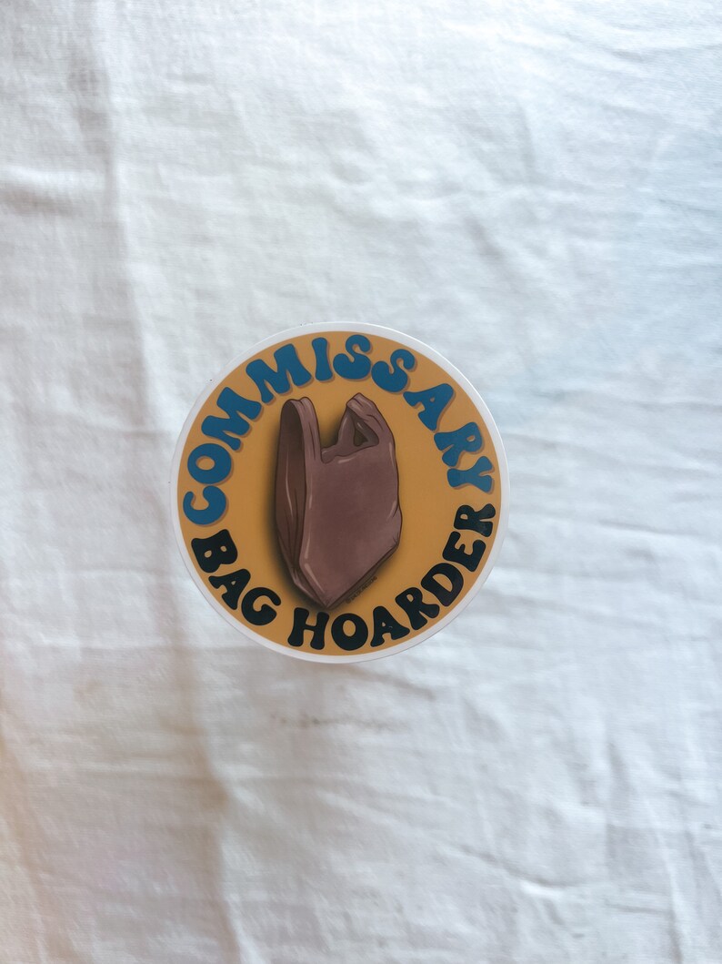 Op de afbeelding: Een ronde sticker met een gele achtergrond en blauwe tekst die "Commissary Bag Hoarder" luidt, met een bruine plastic tas in het midden.
