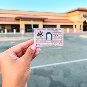May include: A pink and white United States Uniformed Services Identification and Privilege Card. The card features a portrait, text, and the US military emblem. The card is held in front of a building.
