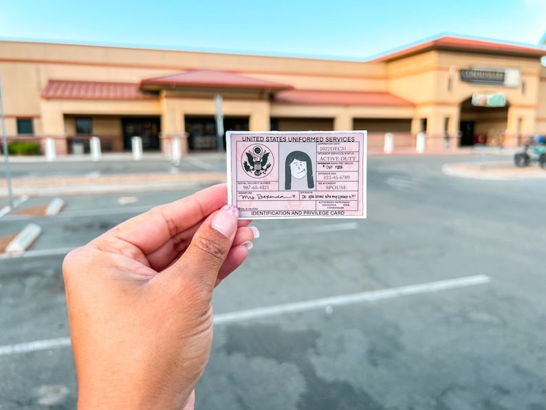 May include: A pink and white United States Uniformed Services Identification and Privilege Card. The card features a portrait, text, and the US military emblem. The card is held in front of a building.