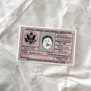 May include: A pink and white sticker designed to look like a United States Uniformed Services ID card. It features a cartoon drawing of a person, the name "Mr. Dependa", and the question "Do you know who my spouse is?!?".