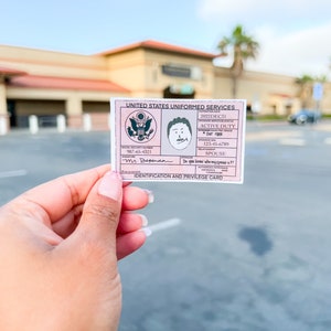 May include: A pink United States Uniformed Services identification and privilege card. The card includes the text "ACTIVE DUTY", "SPOUSE", and "Mr. Dependa". The card also features a cartoon drawing of a man's face and the US Armed Forces seal.