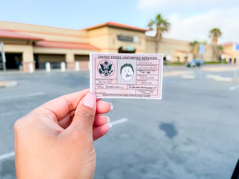 May include: A pink United States Uniformed Services identification and privilege card. The card includes the text "ACTIVE DUTY", "SPOUSE", and "Mr. Dependa". The card also features a cartoon drawing of a man's face and the US Armed Forces seal.