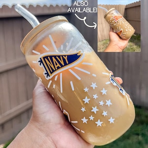 May include: A clear glass tumbler with a bamboo lid and a straw, filled with a light brown beverage. The tumbler features white star and burst designs, along with a "NAVY" banner. A smaller image shows a similar tumbler.