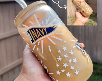 May include: A clear glass tumbler with a bamboo lid and a straw, filled with a light brown beverage. The tumbler features white star and burst designs, along with a "NAVY" banner. A smaller image shows a similar tumbler.