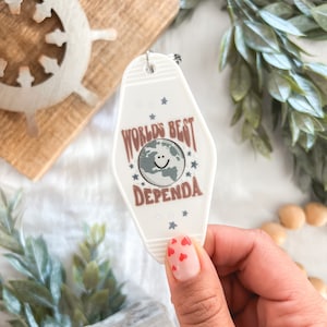 May include: White key chain with a light brown, blue, and white design. The key chain has the text "World's Best Dependa" with a smiley face and stars.