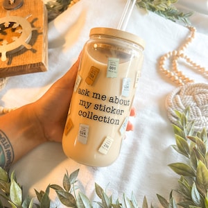 May include: A clear glass tumbler with a bamboo lid and a straw. The tumbler is covered in small stickers with numbers and text that reads "ask me about my sticker collection".