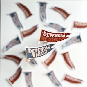 May include: A set of two colorful pennant flags with the text "DependAs: We Serve Too" and "DependAs: Unite!" printed on them. The flags are made of paper and have a distressed, vintage look.