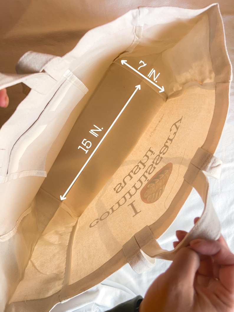 May include: A white canvas tote bag with a brown logo that says "I  comma  love  nature" and a brown leaf design. The bag measures 15 inches wide and 7 inches tall.