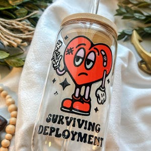 May include: A clear glass tumbler with a bamboo lid and a clear straw. The tumbler features a red heart-shaped cartoon character with a bandage and the words "SURVIVING DEPLOYMENT" in black.