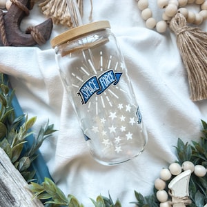 May include: A clear glass tumbler with a bamboo lid and a clear straw. The tumbler features a "SPACE FORCE" design with stars and a blue banner. The design is white and blue. The tumbler is on a white surface.