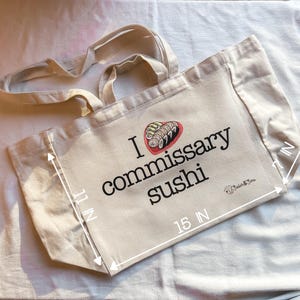 May include: A beige canvas tote bag with black text that reads "I love commissary sushi" and a graphic of sushi rolls. The bag measures 15 inches wide and 11 inches tall.