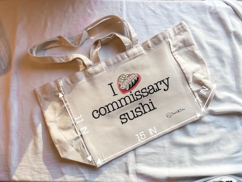 May include: A beige canvas tote bag with black text that reads "I love commissary sushi" and a graphic of sushi rolls. The bag measures 15 inches wide and 11 inches tall.