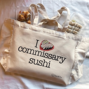 May include: A white canvas tote bag with the text "I love commissary sushi" and a graphic of sushi rolls in a heart shape. The bag has a ruffled edge and two handles.