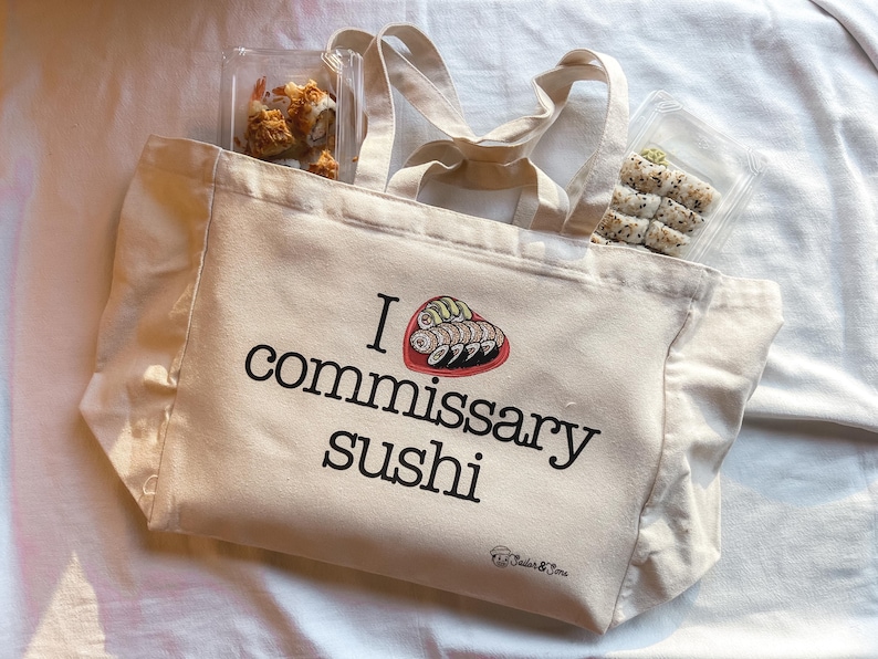 May include: A white canvas tote bag with the text "I love commissary sushi" and a graphic of sushi rolls in a heart shape. The bag has a ruffled edge and two handles.