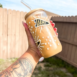 May include: A reusable glass tumbler with a straw, featuring a black and white design with stars and the word "ARMY" in a banner. The tumbler is filled with a light brown beverage.