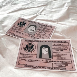 May include: Two pink and grey Identification and Privilege Cards from the United States Uniformed Services. Each card features a cartoon illustration of a person and includes text such as "Active Duty" and "Spouse."
