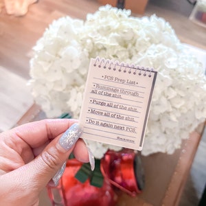 May include: A small notepad with a spiral binding, featuring a "PCS Prep List" with bullet points. The list includes phrases like "Rummage through all of the shit" and "Purge all of the shit." The notepad is held in front of a bouquet of white flowers and a red decorative item.