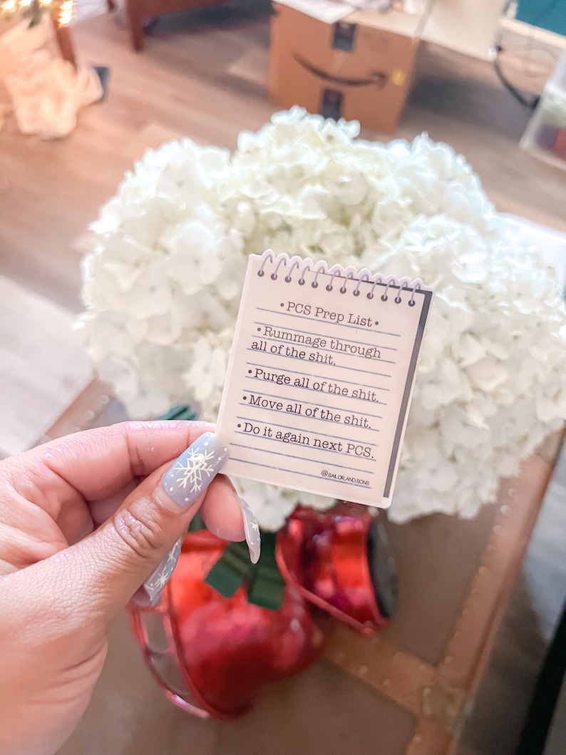 May include: A small notepad with a spiral binding, featuring a "PCS Prep List" with bullet points. The list includes phrases like "Rummage through all of the shit" and "Purge all of the shit." The notepad is held in front of a bouquet of white flowers and a red decorative item.