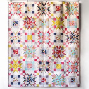 Puede incluir: Una colcha rectangular con un patrón repetitivo de bloques de estrellas multicolores y pequeños bloques cuadrados. La tela muestra varios estampados florales y geométricos en tonos rojo, amarillo, azul, rosa y blanco sobre un fondo claro.