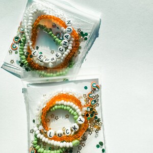 May include: Two sets of colorful beaded bracelets with the words "LEPRECHAUN" and "LUCKY" spelled out in white letters. The bracelets are packaged in clear plastic bags with green, gold, and rainbow confetti.
