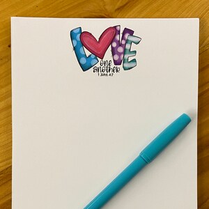 May include: A white notepad with the word "LOVE" in colorful lettering, a blue pen, and the name "amy williams" in blue script. The word "LOVE" has a heart and polka dots.