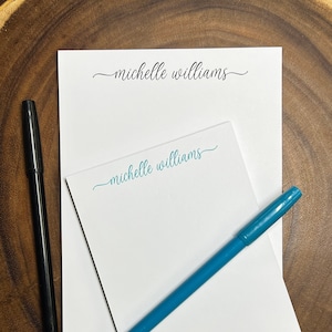 May include: A white notepad with the name "michelle williams" written in cursive on the top and bottom. A blue pen and a black pen are lying on top of the notepad.