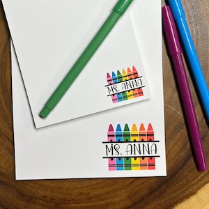 May include: White stationery set with a green pen, a purple pen, and a blue pen. The stationery features a colorful crayon design with the text "Ms. Anna". The set is on a wooden surface.