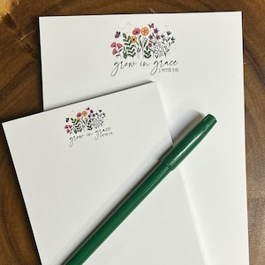 May include: Two white notepads with a floral design and the text "grow in grace 2 Peter 3:18". The smaller notepad has the name "Mrs. Williams" written on it. A green pen is lying on top of the notepads.