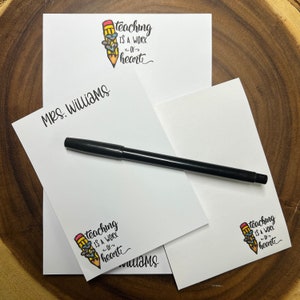 May include: A stack of white notepads with a black pen on top. The notepad has a pencil illustration with the text "teaching is a work of the heart" and the name "Mrs. Williams" printed on the front.