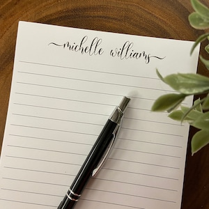 May include: A white lined notepad with the name "michelle williams" in elegant script. A black and silver pen rests on the notepad. The notepad is on a wooden surface.