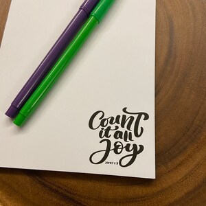 May include: White notepad with the text "Count it all joy" in black cursive lettering. The text "James 1:2" is printed in smaller font below the main text. Two pens, one green and one purple, are lying on top of the notepad.
