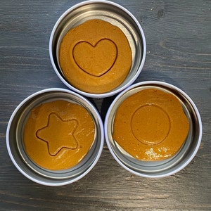 May include: Three round metal containers with lids, each containing a brown, caramel-like candy with a different shape cut out of the center. The shapes are a heart, a star, and a circle.