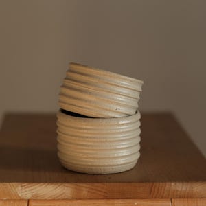 May include: Two stacked, off-white ceramic bowls with horizontal ridged detailing. The bowls are set on a wooden surface, with a neutral background. The top bowl is slightly tilted, revealing the dark interior of the bottom bowl.
