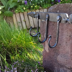 Puede incluir: Gancho de pared de metal forjado a mano con tres ganchos curvos. El gancho está montado en un pilar de piedra en un entorno de jardín. El metal es de color gris oscuro y la piedra es de color marrón rojizo. El gancho tiene un extremo puntiagudo decorativo.
