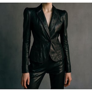 Puede incluir: Blazer de cuero negro con cierre de un solo botón, combinado con pantalones de cuero a juego. La chaqueta presenta solapas con muescas y un ligero acolchado en los hombros. El conjunto se presenta sobre un fondo gris neutro, resaltando la silueta elegante y entallada.