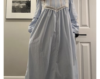 Puede incluir: Un camisón azul claro de manga larga con cuello alto y canesú con ribete de encaje blanco. El camisón es largo y tiene bolsillos. Un cuadro de paisaje en blanco y negro enmarcado cuelga de la pared.